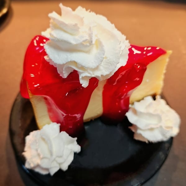 A slice of cheesecake with bright red cherry topping and three dollops of whipped cream.