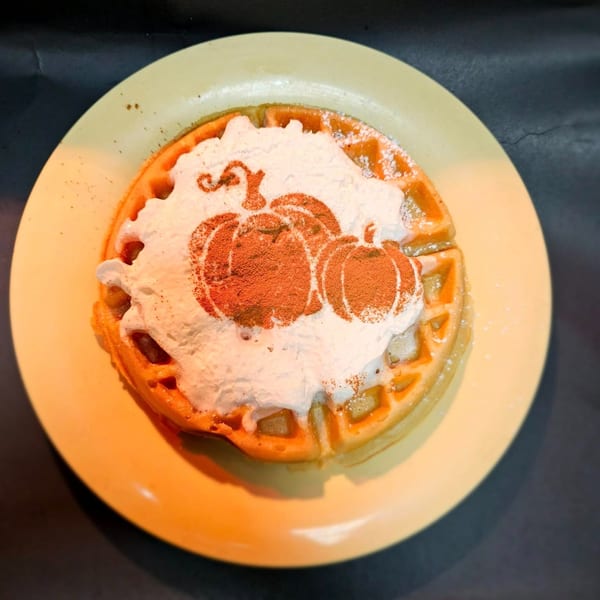A waffle topped with whipped cream and a pumpkin spice design on a yellow plate.