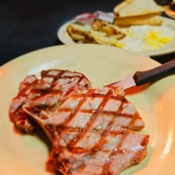 Grilled pork chops with a knife on a yellow plate, with eggs, potatoes, and toast in the background.